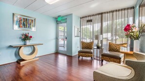 A hotel lobby with a wooden floor, a framed abstract painting, a vase with flowers, and a chair with a patterned cushion. - Rodeway Inn Anderson North - Clemson (Anderson)