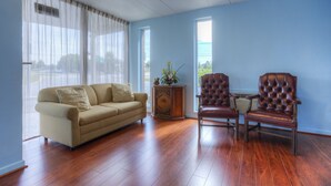 A living room with a beige sofa, two leather armchairs, a wooden side table with a vase, and large windows with curtains. - Rodeway Inn Anderson North - Clemson (Anderson)