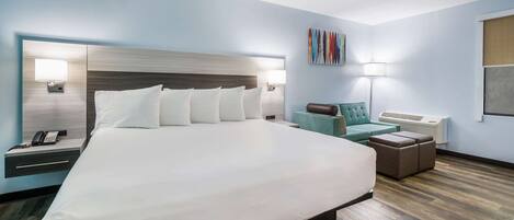 A modern hotel room with a large bed, a desk with a phone, a lamp, and a seating area with a chair and ottoman.