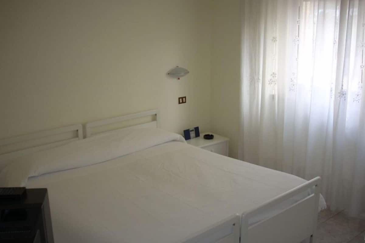 A single-bed room with a white bed, a bedside table with a lamp, and a window with sheer curtains.