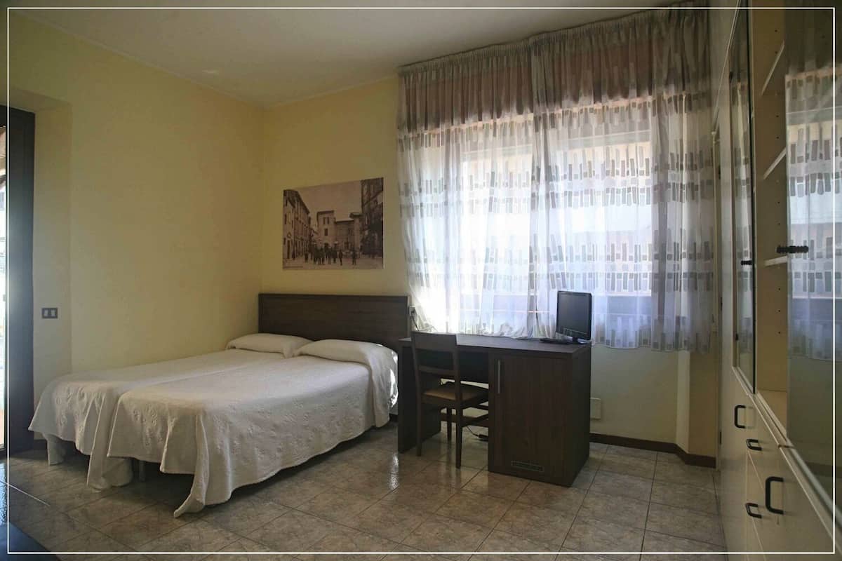 A bedroom with a bed, a desk, and a chair. There is a window with curtains and a painting on the wall.