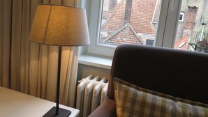 Una habitación con ventana que da a un edificio histórico con tejados de tejas rojas. Una lámpara está sobre una mesa junto a la ventana, y se ve un radiador debajo de la ventana.