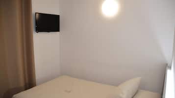 Ein Einzelbett mit weißer Matratze und Kissen, ein flachbildfernseher an der Wand montiert und eine Leuchte über dem Bett.