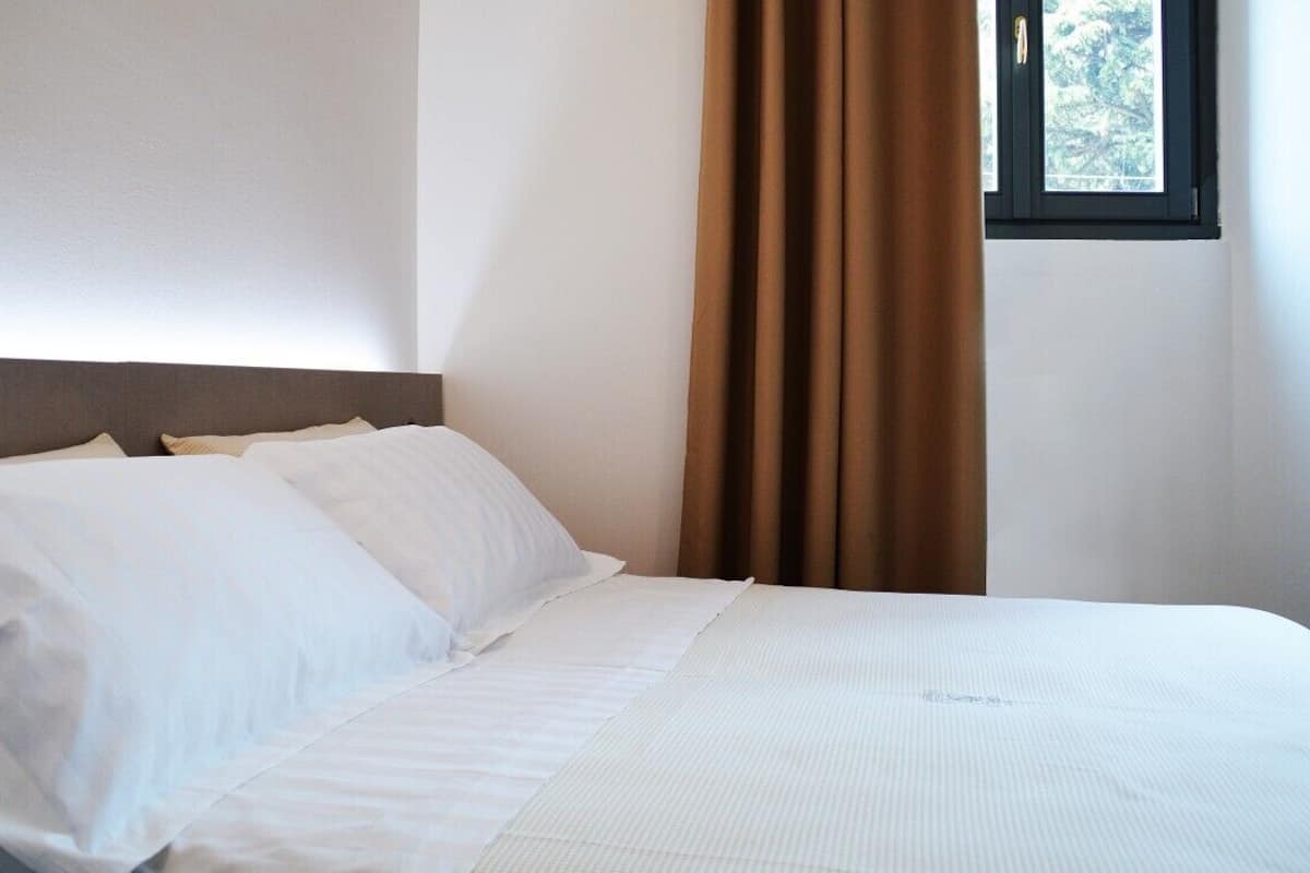 Una cama bien hecha con sábanas blancas, cabecera y ventana con cortinas marrones.