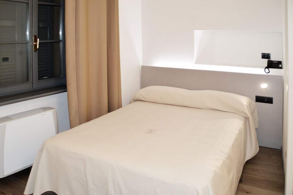 Un dormitorio pequeño y ordenado con una cama individual, una ventana con cortinas, un radiador y una lámpara fijada a la pared.