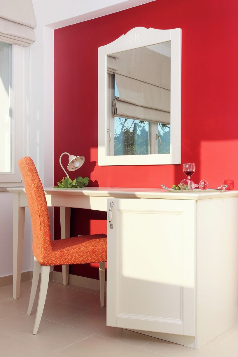 Una habitación con pared roja, un escritorio blanco, una silla blanca, un armario blanco, una lámpara blanca y una copa de vino.
