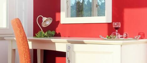 Una habitación con pared roja, un escritorio blanco, una silla blanca, un armario blanco, una lámpara blanca y una copa de vino.