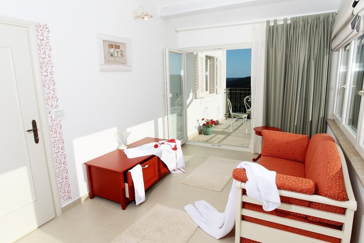 A room with a red sofa, a white armchair, a small table, and a glass door leading to a balcony with a view of the sea.