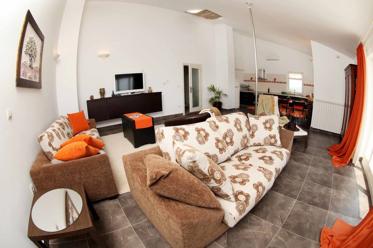 A modern living room with a sofa, a coffee table, and a TV. There is a dining area with a table and chairs, and a kitchen in the background.