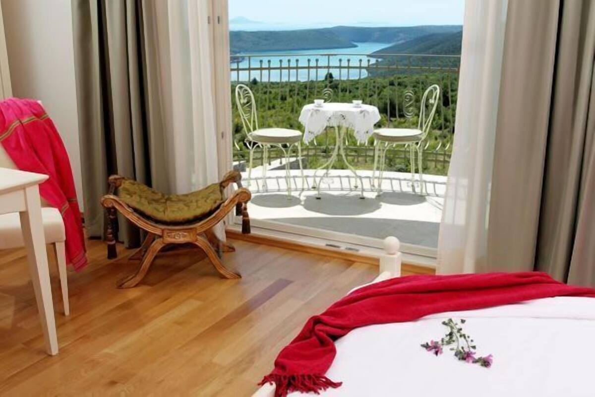 A bedroom with a balcony, a wooden chair, a small table, and a view of a lake.