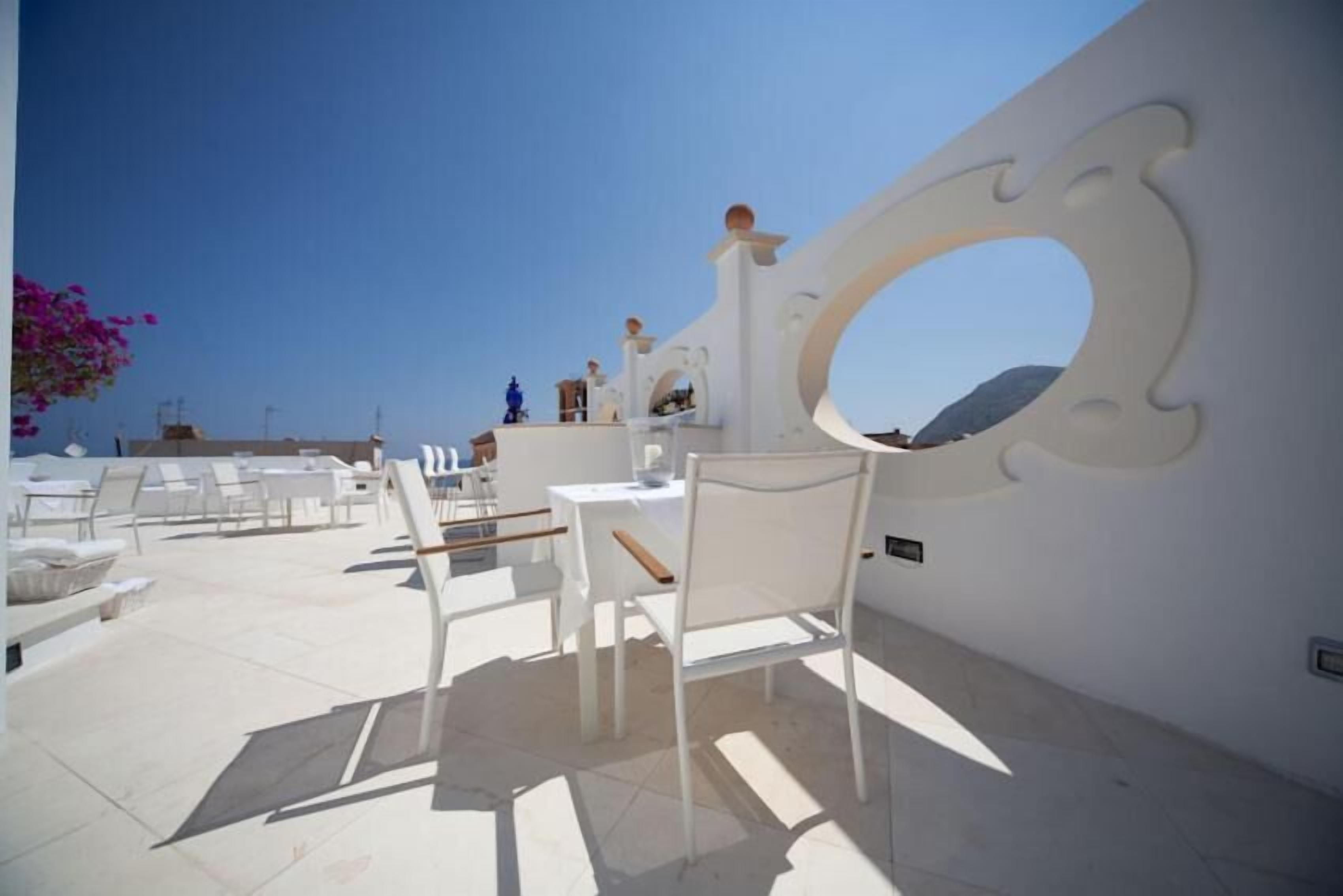 Area esterna con sedute e tavoli bianchi, parete decorativa con un’apertura circolare e vista libera sul cielo.