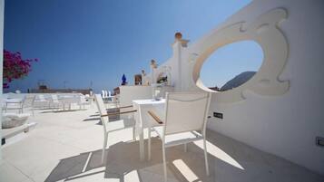 Area esterna con sedute e tavoli bianchi, parete decorativa con un’apertura circolare e vista libera sul cielo.