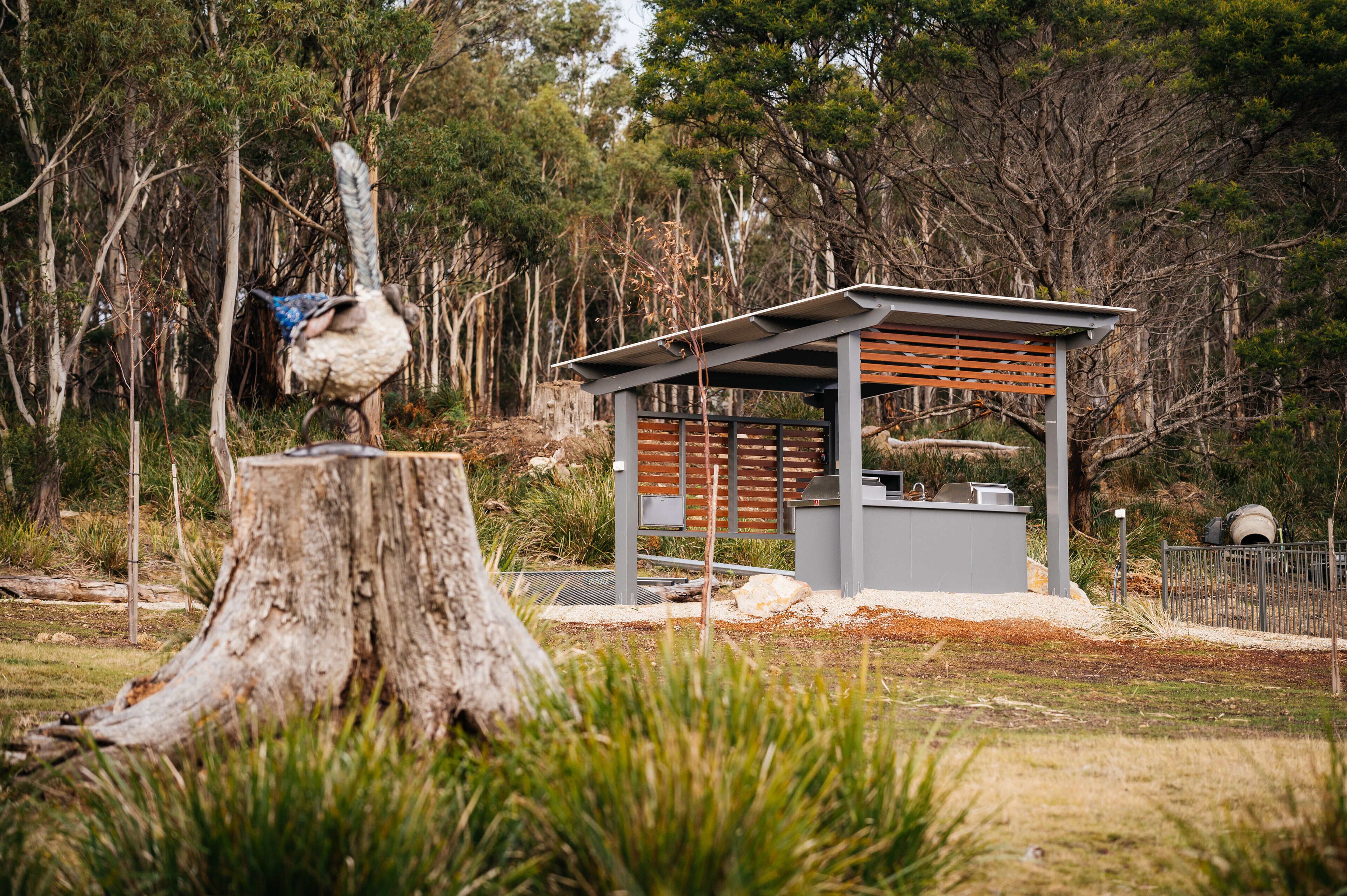 A small pavilion with a slatted roof and a bench, surrounded by trees and a large tree stump.