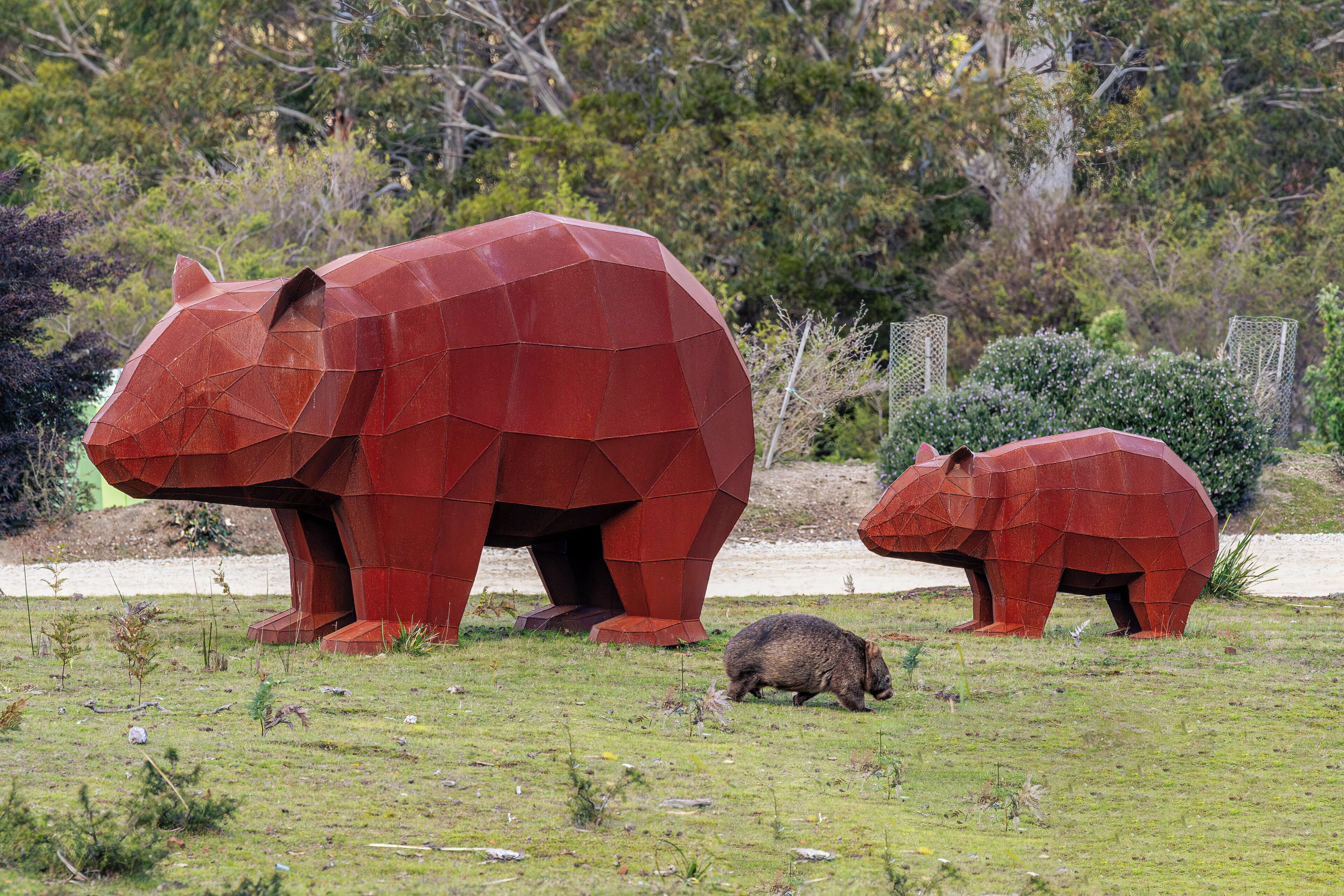 Una gran escultura metálica roja de un oso en un campo cubierto de hierba, con dos osos más cerca, uno de ellos marrón.