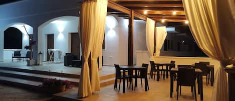 An outdoor patio area with tables and chairs, covered by a wooden pergola and draped curtains.