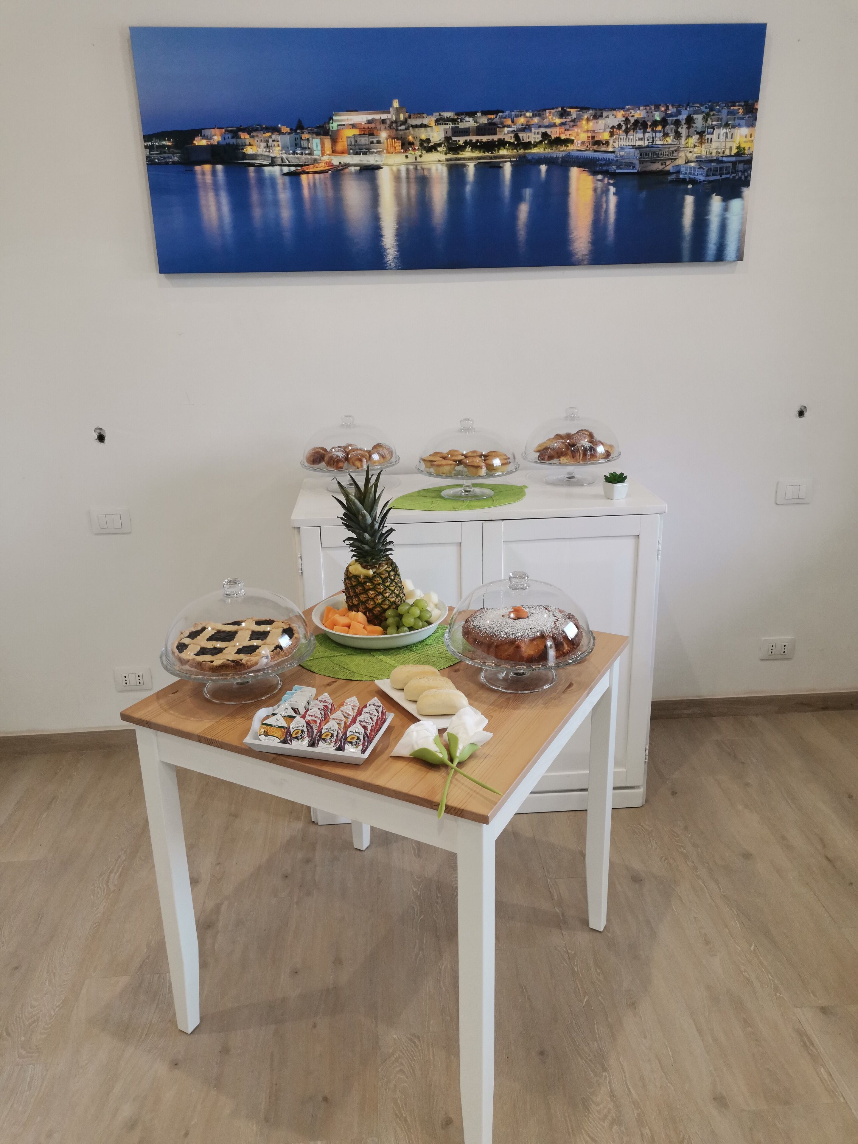A dining table with various desserts and a pineapple centerpiece, set against a white wall with a cityscape painting.