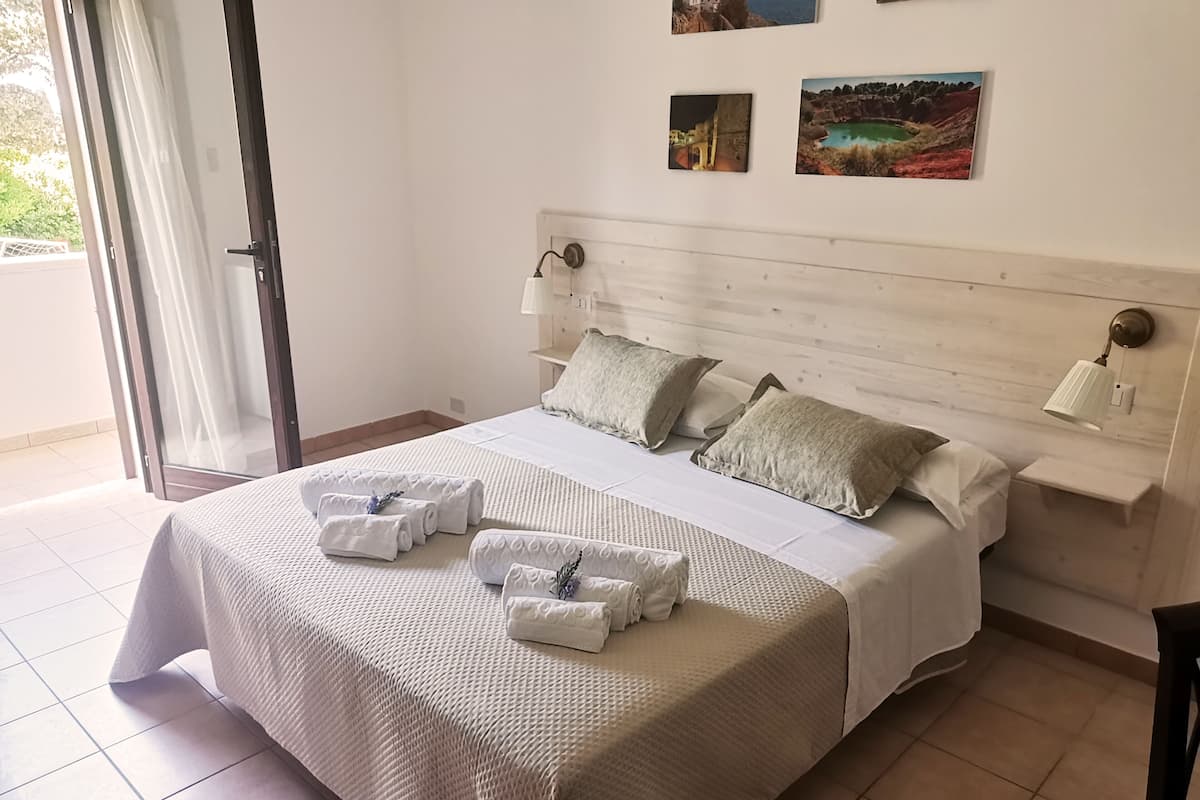 A neatly made bed with towels, a headboard with wall-mounted lamps, and a view of the outdoors through a sliding door.