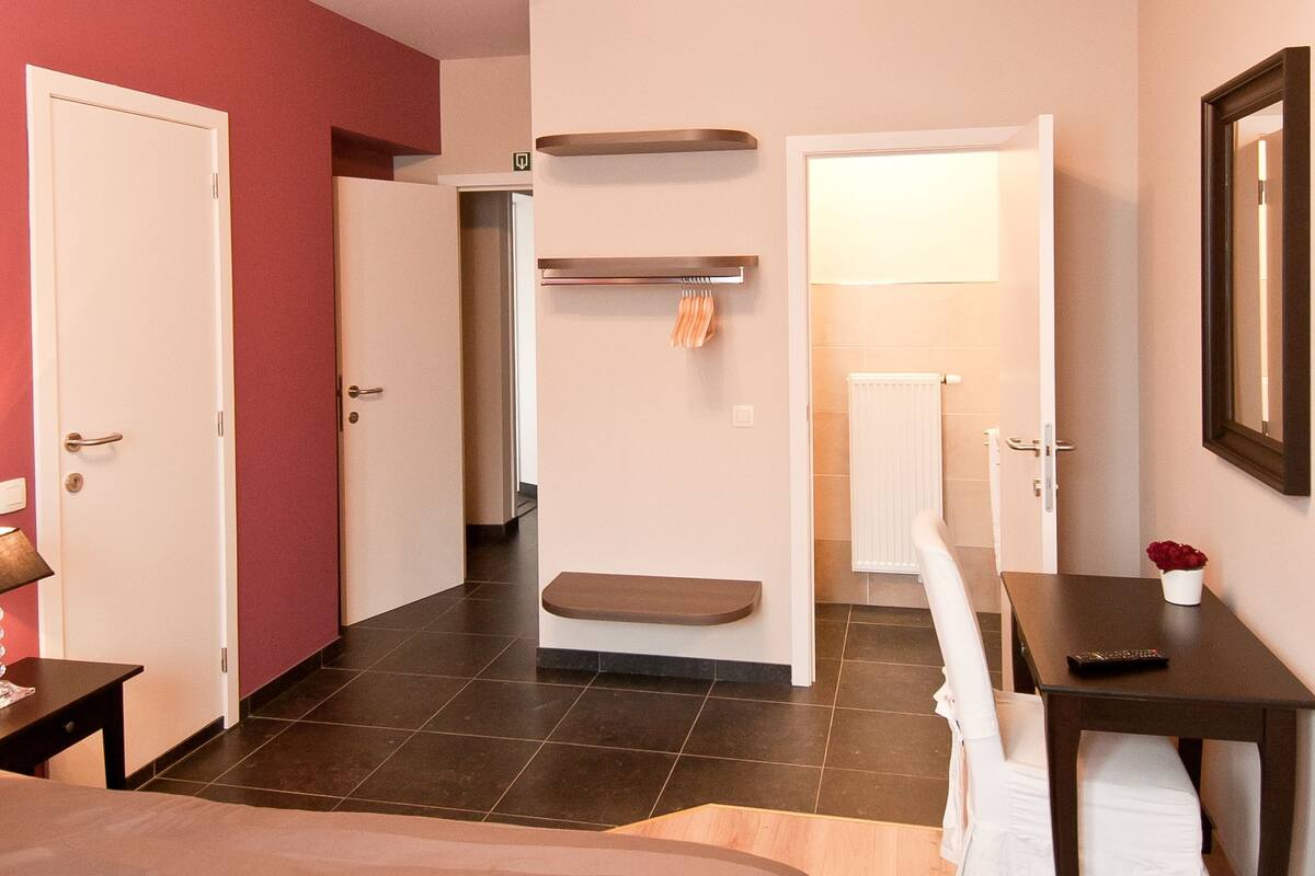 A hotel room with a bed, a desk, a chair, a bench, a coat rack, and a bathroom visible through an open door.
