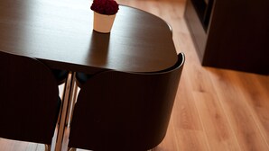 Un comedor con una mesa y sillas, una rosa roja sobre la mesa, una ventana con luz natural, un radiador y un mueble oscuro con una planta.