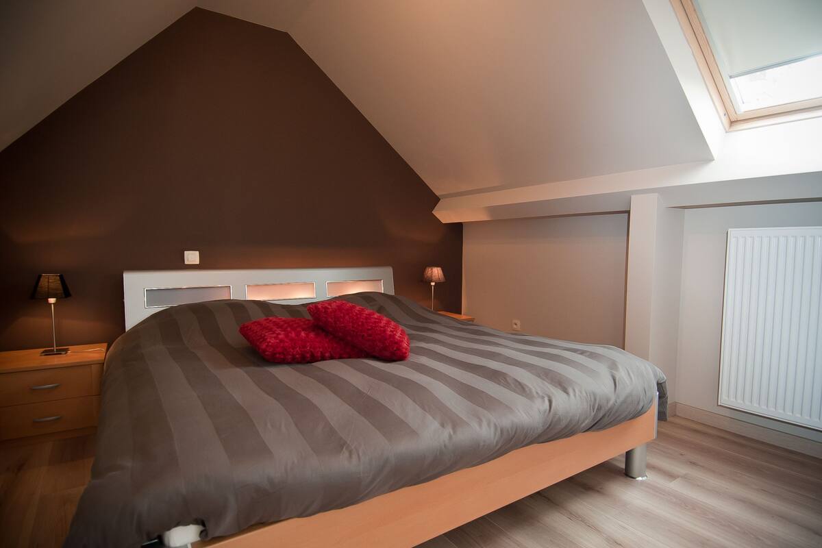 A bedroom with a sloped ceiling, a large bed with a striped bedspread, two red pillows, a bedside table with a lamp, and a window with blinds.