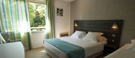 Habitación de hotel con cama, escritorio, silla, ventana con cortinas y lámpara de pared.