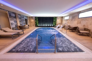 Una piscina interior moderna con sillas de descanso y un fondo de pared de piedra.