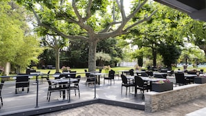 Un área de comedor al aire libre con mesas y sillas, rodeada de árboles y un muro de piedra.
