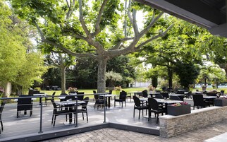 Un área de comedor al aire libre con mesas y sillas, rodeada de árboles y un muro de piedra.