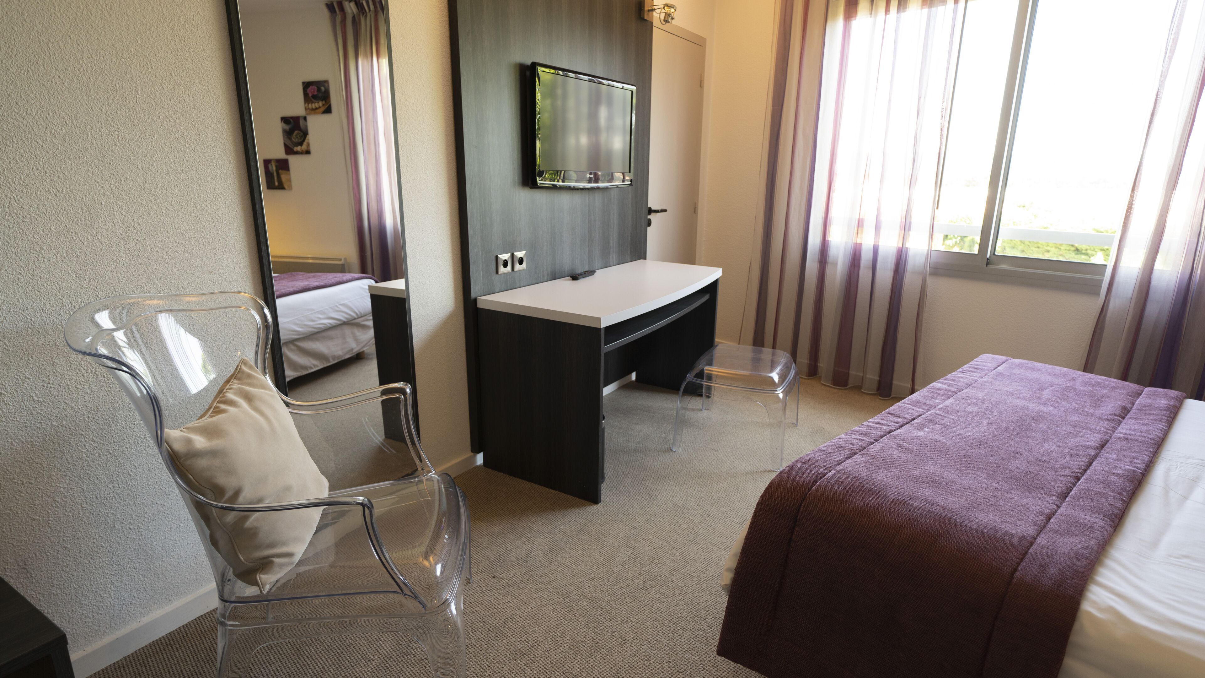 A hotel room with a bed, a desk, a chair, a mirror, and a window with curtains.