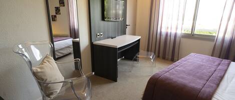 A hotel room with a bed, a desk, a chair, a mirror, and a window with curtains.