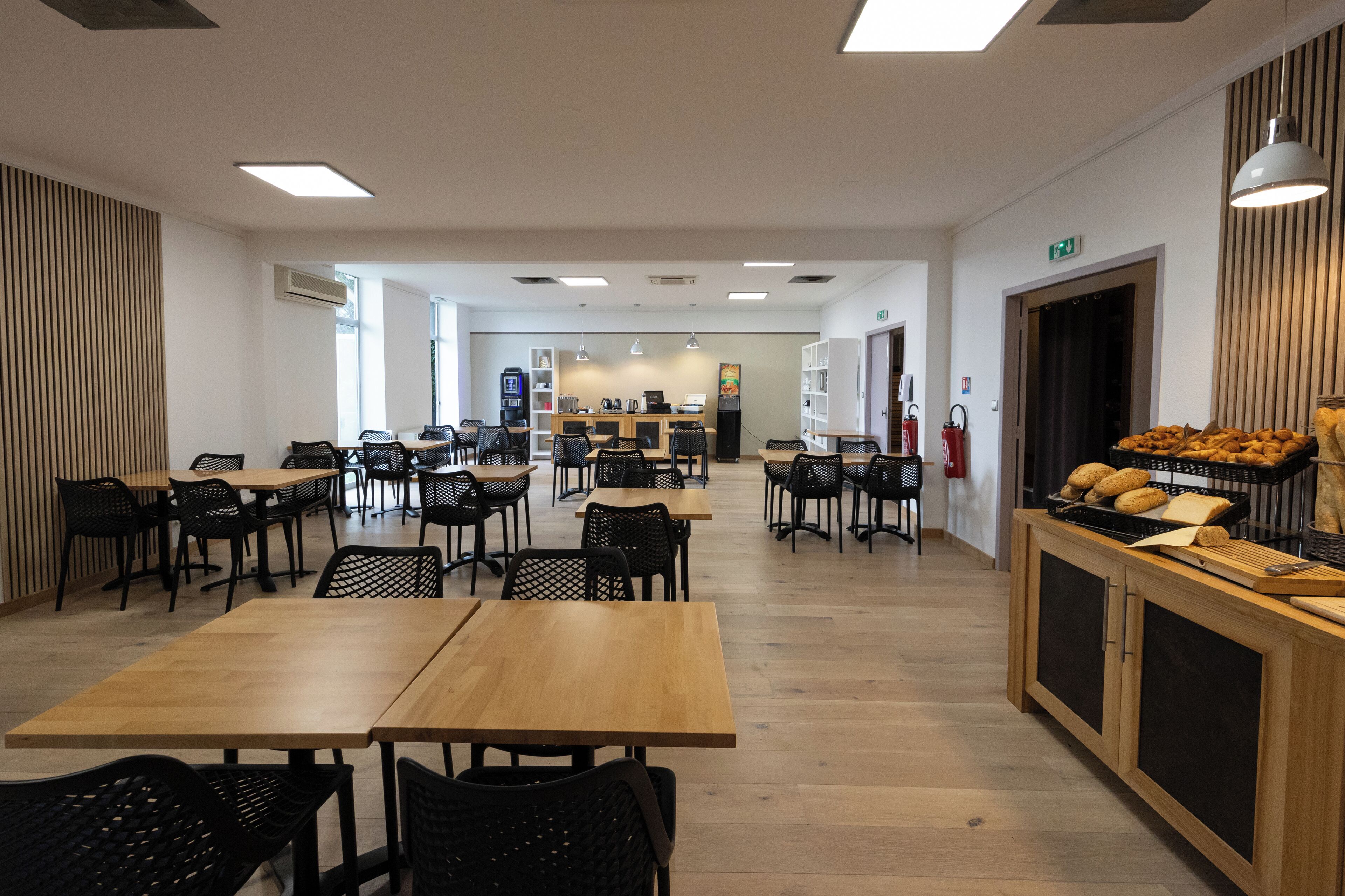 Una amplia cafetería con varias mesas y sillas, un mostrador con productos de panadería y un letrero de salida visible.