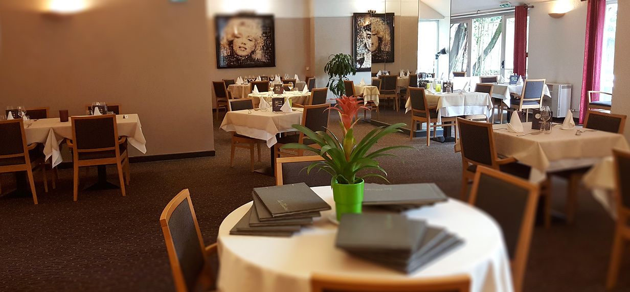Comedor de hotel con varias mesas y sillas, algunas con manteles, y una planta en maceta sobre una de las mesas.