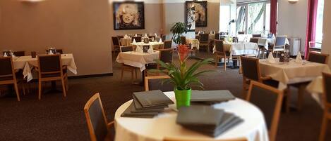 Comedor de hotel con varias mesas y sillas, algunas con manteles, y una planta en maceta sobre una de las mesas.