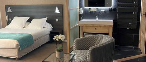 A modern hotel room with a bed, a sitting area with two chairs, a small table with a vase, a vanity with a mirror, and a black tiled wall.