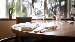 Mesa de comedor dispuesta con servilletas blancas, cubiertos y copas de vino, con vista a los árboles a través de la ventana.