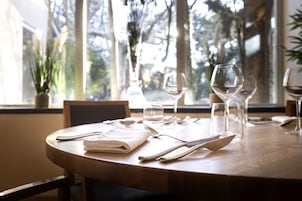 Mesa de comedor dispuesta con servilletas blancas, cubiertos y copas de vino, con vista a los árboles a través de la ventana.