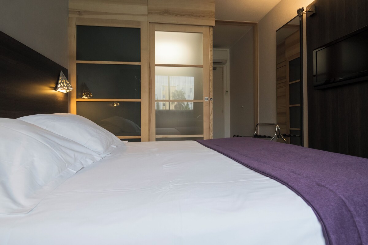 A bedroom with a bed, a wardrobe, a TV, and a window with a view of buildings.