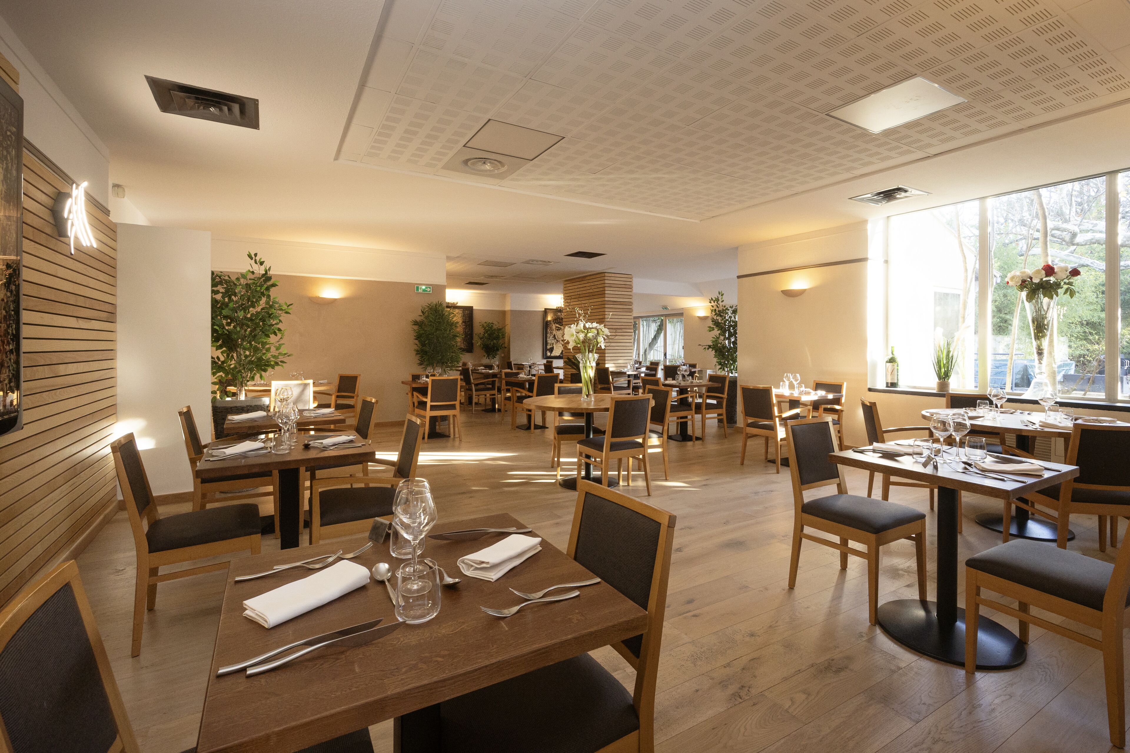 A spacious restaurant with multiple dining tables and chairs, large windows, and indoor plants.