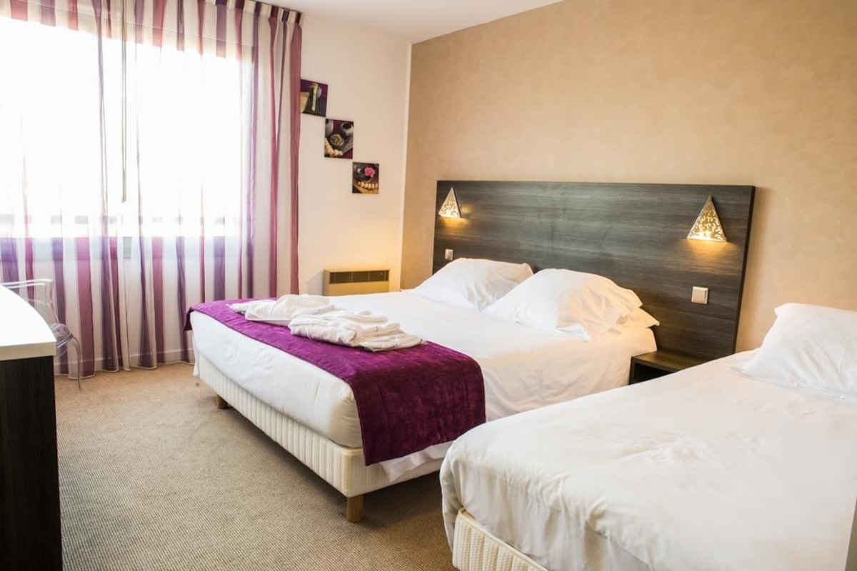 A hotel room with two beds, a large window with curtains, a headboard with wall-mounted lights, and a small bedside table.