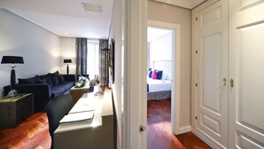 A modern living room with a sofa, coffee table, and lamps. A bedroom with a bed and curtains is visible through an open door. - Boutike Cibeles (Madrid)