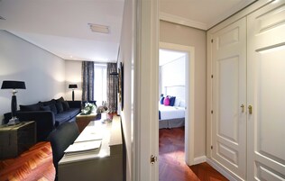 Una sala de estar moderna con un sofá, una mesa de centro y lámparas. Se ve un dormitorio con una cama y cortinas a través de una puerta abierta.