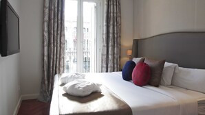 A neatly made bed with a white comforter and pillows, a bedside table with a lamp, and a window with curtains. - Boutike Cibeles (Madrid)
