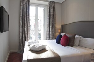 Una cama bien hecha con edredón blanco y almohadas, una mesita de noche con lámpara y una ventana con cortinas.