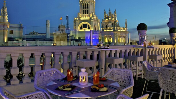 An outdoor dining area with tables and chairs, a view of a historic building with a clock tower, and a railing with decorative elements. - Boutike Cibeles (Madrid)