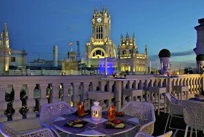 An outdoor dining area with tables and chairs, a view of a historic building with a clock tower, and a railing with decorative elements. - Boutike Cibeles (Madrid)