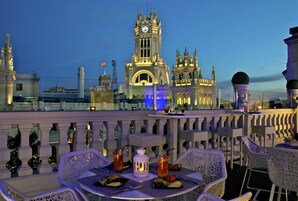Un área de comedor al aire libre con mesas y sillas, vistas a un edificio histórico con una torre de reloj, y una barandilla con detalles decorativos.