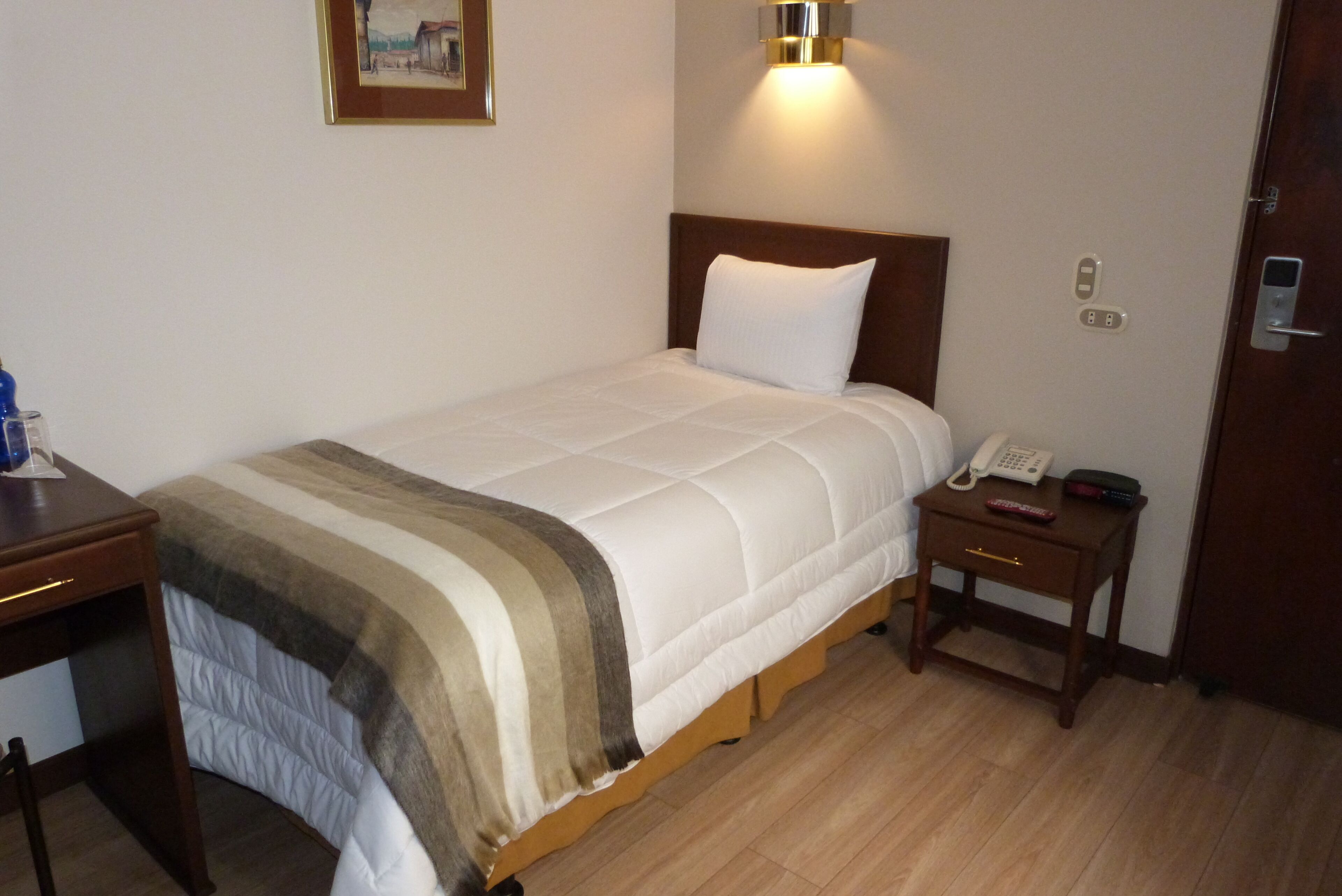 Una cama bien tendida con una colcha a rayas, una mesita de noche con un teléfono y control remoto, y una lámpara de pared.