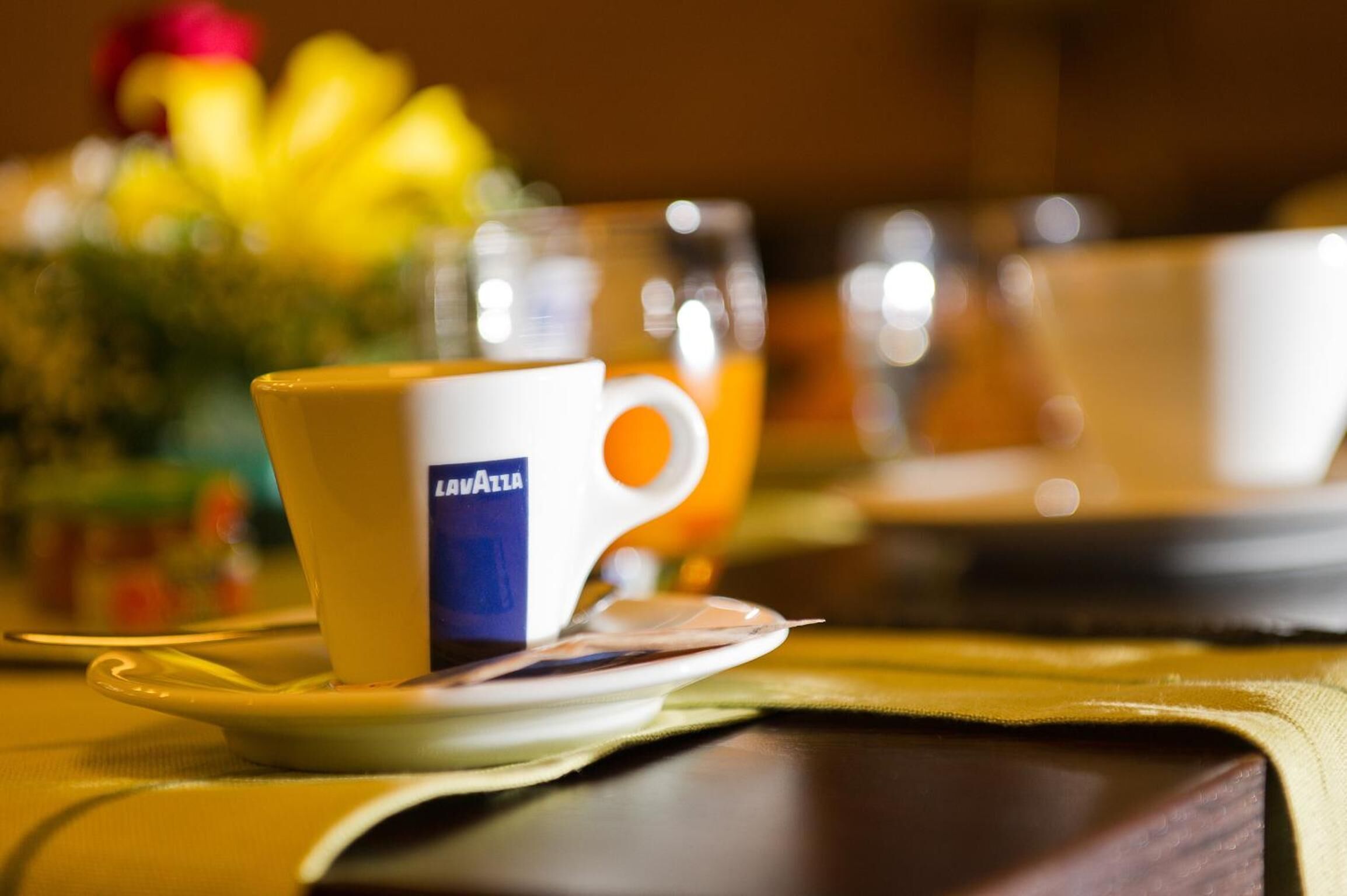 Una taza con un logo sobre un platito, un vaso de jugo de naranja y un fondo borroso con una flor amarilla.