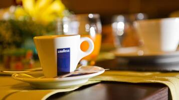 Una taza con un logo sobre un platito, un vaso de jugo de naranja y un fondo borroso con una flor amarilla.