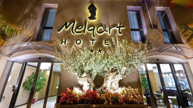 La façade de l’hôtel Melqart, avec une enseigne bien visible, un grand arbre et un parterre de fleurs devant l’entrée.